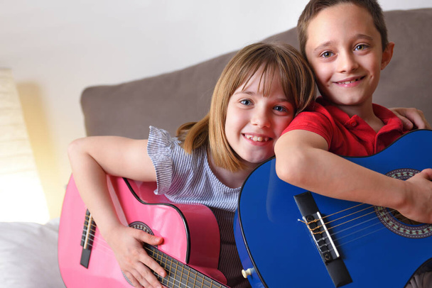 Little girl and boy caught and smiling practicing the guitar sitting on a sofa in their living room - Photo, Image