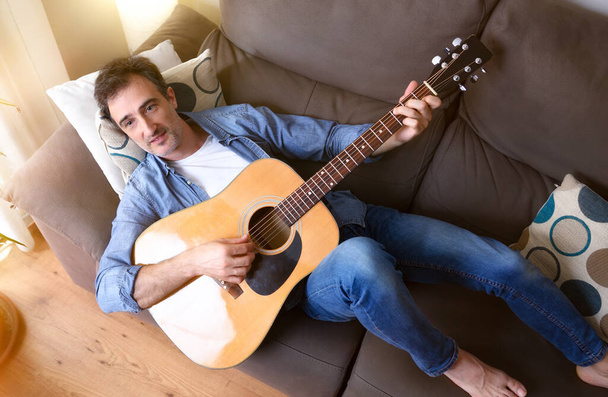 Detail relaxed and smiling man lying on a sofa playing the guitar in a room at home. Top view. - Foto, imagen
