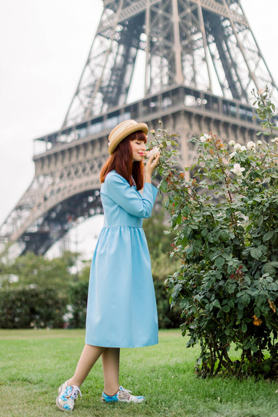 Young beautiful woman, dressed in elegant light blue dress and straw hat, walks on the Paris city street near the flowering rose bushes, touching and smelling the flowers. Eiffel tower on background - Foto, imagen