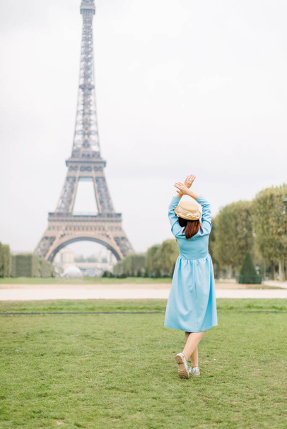 Back view of pretty lady in blue dress and hat, enjoying walk in Paris, near Eiffel tower. Young attractive happy woman with hands up facing the Eiffel Tower in Paris, France. - Valokuva, kuva