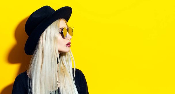 Студийный портрет молодой блондинки в черной шляпе и солнцезащитных очках. Отводя взгляд на пустой фон желтого цвета с копировальным пространством
.  - Фото, изображение