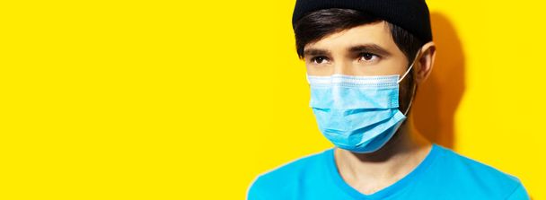 Studioportret van een jonge hipster met medisch griepmasker, bescherming tegen het coronavirus. Covid-19 preventie. Panoramisch bannerzicht. achtergrond van gele kleur met kopieerruimte. - Foto, afbeelding