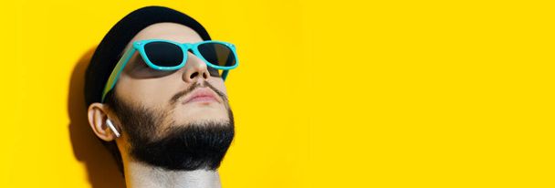 Panoramic banner view, portrait of young hipster, looking up, using wireless earphones, wearing shades of aqua menthe color on background of yellow with copy space. - Zdjęcie, obraz