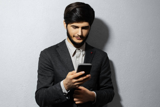 Studio portrait of young confident businessman wearing suit, holding and using smartphone. Background of grey textured wall. - Foto, Imagen