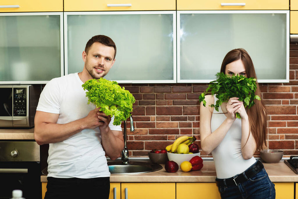 Sağlıklı gıda reklamı. Çekici erkek ve kadın ellerinde yeşilliklerle moda bir mutfakta poz veriyorlar. Kadın yüzünün yarısını bir demet dereotu ve maydanozla kaplamış. - Fotoğraf, Görsel