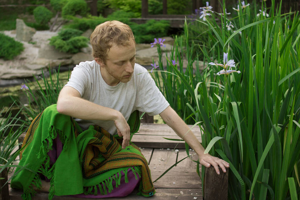Homem bonito jovem senta-se em uma ponte de madeira na grama e alcança uma flor
 - Foto, Imagem