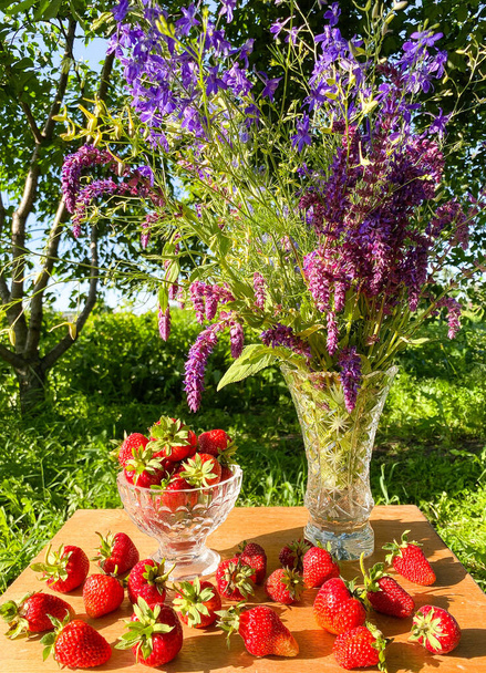 Un gran ramo de flores silvestres de colores en un jarrón de vidrio y fresas rojas maduras en una superficie de madera abstracta. Composición sobre el fondo de plantas verdes en el jardín. Espacio libre. Desenfocar fondo claro
. - Foto, imagen