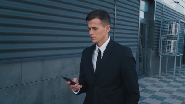 Side view of confident businessman in suit walking with phone. Man looking into smartphone outdoors - Πλάνα, βίντεο
