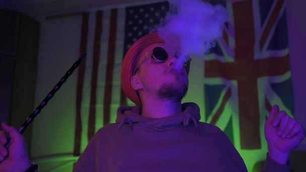 Stylish hipster smokes a hookah in home interior against backdrop of American and Great Britain flags hanging on the wall - Fotó, kép
