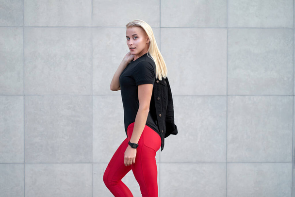 Portrait of young attractive sporty girl or model wearing black sportswear, trendy red leggings. Outdoor shot on grey wall background.                               - Fotografie, Obrázek