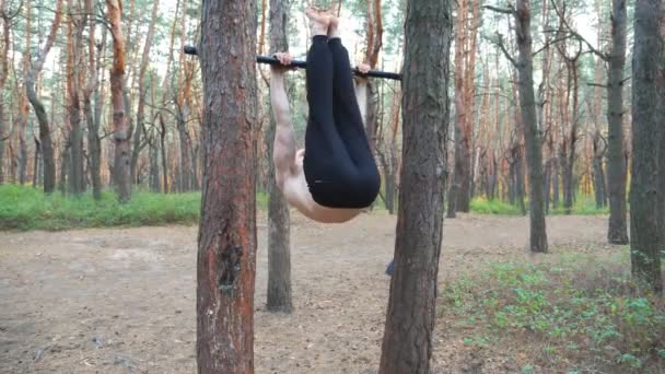 Atletický muž trénuje břišní svaly zvedající nohy na vodorovné tyči. Svalnatý chlap cvičí v lese. Sportovec cvičí venku. Pojetí sportu a aktivního životního stylu. Zpomalený pohyb - Záběry, video