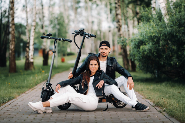 Porträt eines Paares, das in der Nähe von Elektrorollern sitzt und die Zeit in der Natur zusammen genießt, zwei Liebende auf Elektrorollern.. - Foto, Bild