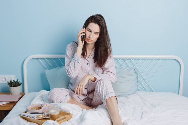 Front view portrait of attractive lovely brunette friendly cheerful girl sitting on bed talking on phone drinking coffe  - Valokuva, kuva