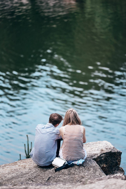 Der Kerl und das Mädchen sitzen auf einer Klippe über dem Fluss und schauen nach vorne. Rückansicht. Liebespaar. vertikales Bild. - Foto, Bild