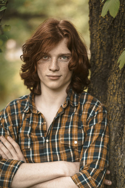 Confident man with long red hair stands with crossed arms leaning against a tree trunk. Sexy Caucasian guy wearing in checkered shirt posing while looking at camera - Foto, Bild