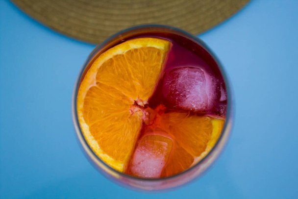 Rotes Getränk auf hellblauem Hintergrund, erfrischende Martini im Glasbecher mit Orangen und Eis auf einem Tisch im Freien mit Strohhut im Hintergrund. - Foto, Bild