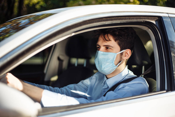 Young man sits behind the steering wheel in the car wearing sterile medical mask. Boy taxi driver works hard during coronavirus outbreak. Social distance, virus spread prevention and treat concept - Foto, imagen