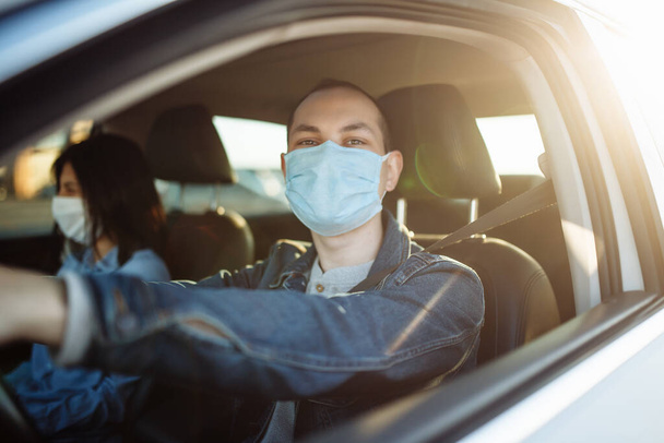 Young man drives a car with a passenger during coronavirus pandemic quaranteene. Taxi driver wears a mask sitiing behind the steering wheel. Social distance, epidemic, protection, health concept - Photo, Image
