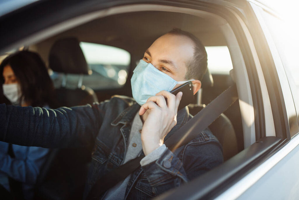 Taxi driver talking on the cell phone and wearing sterile medical mask while waiting in a traffic during coronavirus pandemic. Social distance, new normal, health concept - Photo, Image
