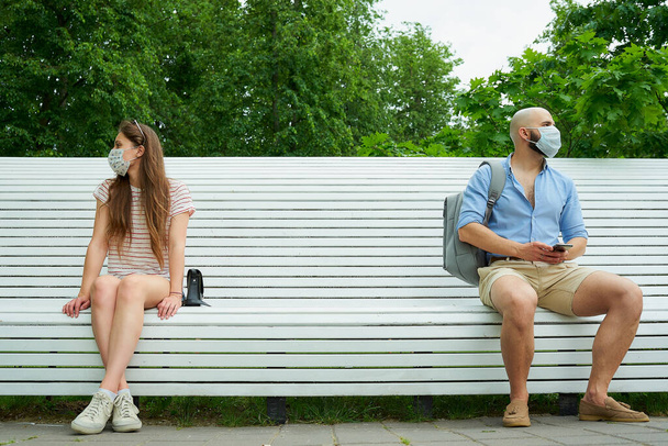Мужчина и женщина смотрят в разных направлениях, сидя на противоположных концах скамейки, держа дистанцию друг от друга, чтобы избежать распространения коронавируса. Парень и девушка в масках на социальное дистанцирование
 - Фото, изображение