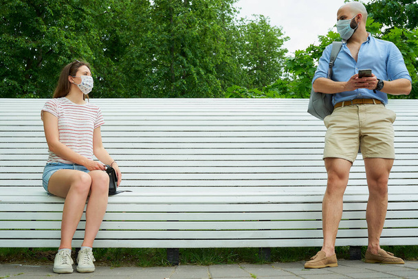 Un hombre esperando cerca de un banco manteniendo una distancia social de una mujer que se sienta en un banco para evitar la propagación del coronavirus. Un chico y una chica con mascarillas quirúrgicas en el distanciamiento social en el parque
. - Foto, imagen