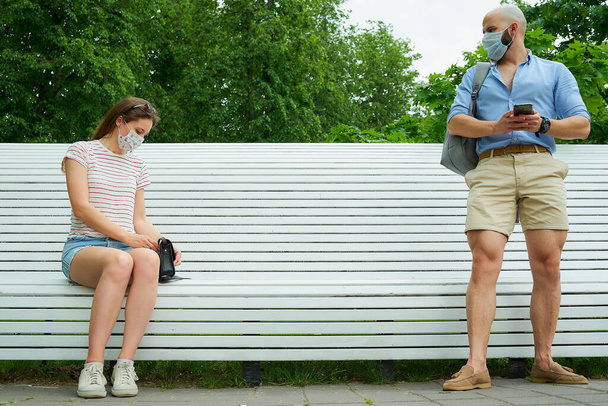 Ένας άντρας που περιμένει κοντά σε ένα παγκάκι κρατώντας μια κοινωνική απόσταση από μια γυναίκα που κάθεται σε ένα παγκάκι για να αποφύγει την εξάπλωση του κορωναϊού. Ένας τύπος και ένα κορίτσι με ιατρικές μάσκες προσώπου σε κοινωνική απόσταση στο πάρκο. - Φωτογραφία, εικόνα