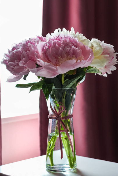 Pink and white large peonies in a glass vase opposite a window with purple curtains. - Foto, Imagen