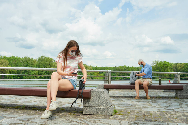 Kobieta na ławce, trzymająca dystans społeczny z łysym mężczyzną siedzącym na innej ławce na promenadzie. Dziewczyna i facet w maskach medycznych odpoczywają w towarzystwie, oddalając się od rzeki.. - Zdjęcie, obraz