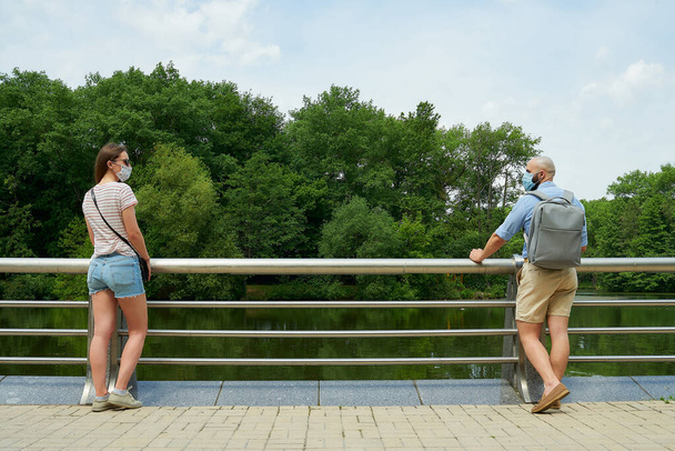 Eine Frau und ein Mann, die einander zuschauen und soziale Distanz wahren, um die Ausbreitung des Coronavirus zu verhindern. Ein Mann und ein Mädchen mit Gesichtsmasken lehnen an den Zaun auf der Promenade - Foto, Bild