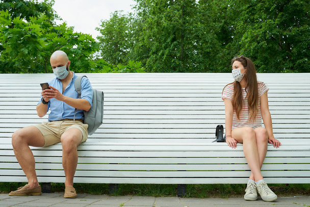 Man met telefoon en jonge vrouw aan weerszijden van een bank die afstand houden van elkaar om de verspreiding van het coronavirus te voorkomen. Guy en een meisje in het gezicht maskers in sociale afstand in het park - Foto, afbeelding