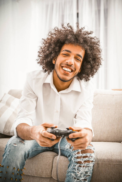 Cute gamer plays game console while sitting on couch. Charming shaggy Arab in ripped jeans and a white shirt toothy smiles enjoying the game. Front view. Toned image - Foto, imagen