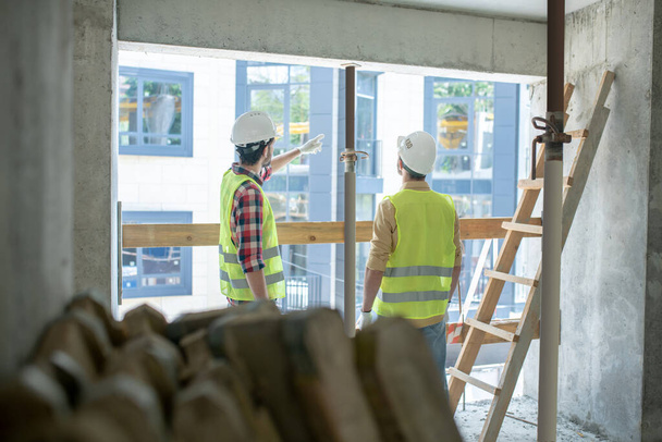 Trabalhadores da construção em coletes amarelos e luvas de pé de volta para a câmera perto da janela, discutindo, apontando para algo
 - Foto, Imagem