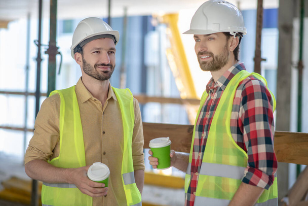 Building workers in yellow vests and helmets having coffee break - Foto, afbeelding
