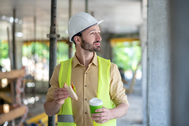 Building worker in yellow vest and helmet holding coffee cup and sandwich - Φωτογραφία, εικόνα
