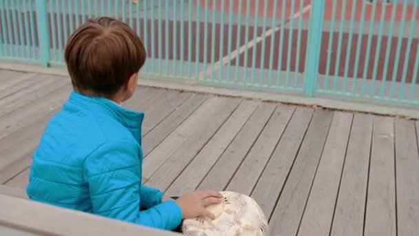 Een kleine jongen die op de bank zit en gedefleerde bal vasthoudt en andere kinderen ziet voetballen. - Video