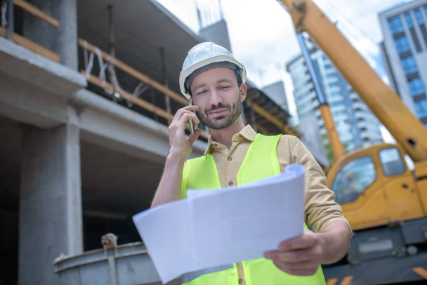 Building worker in helmet and yellow vest holding blueprint, talking on the phone - Φωτογραφία, εικόνα
