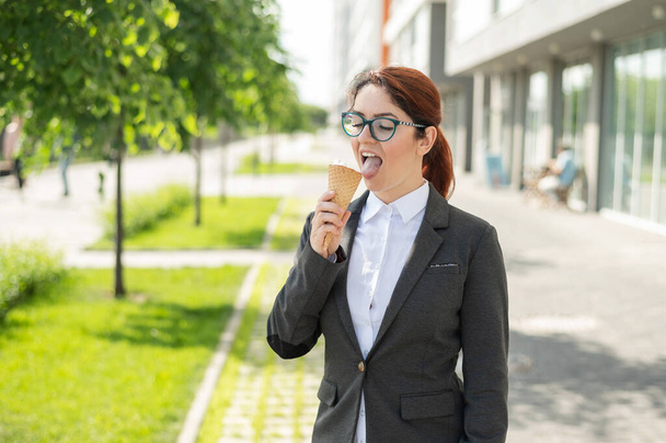 La mujer de negocios está caminando al aire libre y comiendo helado de vainilla en un día soleado de verano. Retrato de una chica sonriente con una camisa blanca y una chaqueta con un cono de helado. Pausa para el almuerzo de un empleado de oficina
. - Foto, Imagen
