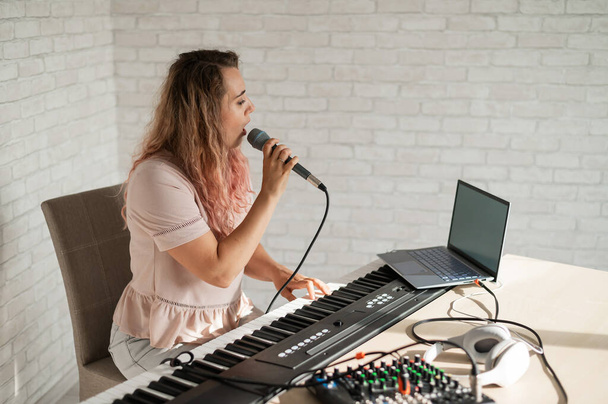 A woman records a vocal lesson using a laptop and accompanying on a keyboard while at home. The teacher sings a song into the microphone and plays the electronic piano. A blogger is recording a video. - Valokuva, kuva