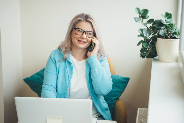 Blonde senior caucasian woman with eyeglasses talking on phone while working remotely at the computer - Photo, Image