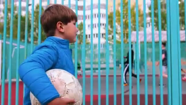 Ein kleiner Junge hält einen Fußballball in der Hand und läuft am Spielplatz vorbei - Filmmaterial, Video