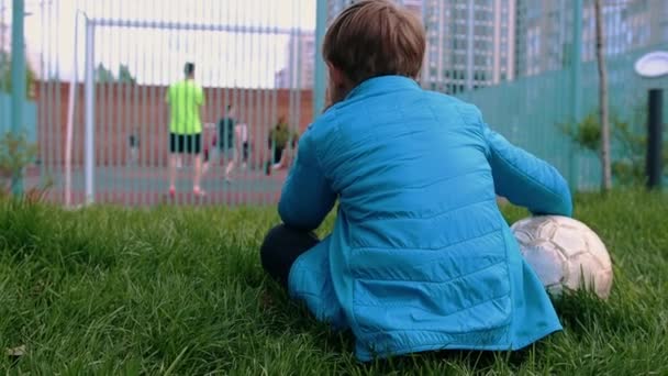 Mały chłopiec siedzący na trawie z piłką nożną i obserwujący innych ludzi grających - Materiał filmowy, wideo