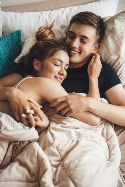 Bájos kaukázusi pár feküdt az ágyban, és ölelkezve reggel mosolyogva csukott szemmel - Fotó, kép