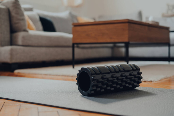Primer plano de un rodillo de espuma negro en una alfombra de fitness, interior de una sala de estar moderna, enfoque superficial selectivo
. - Foto, imagen