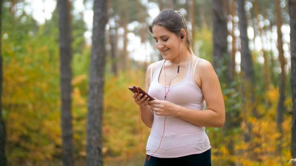 Портрет улыбающейся спортивной женщины, выбирающей музыкальный трек на смартфоне перед пробежкой или бегом в лесу
 - Фото, изображение