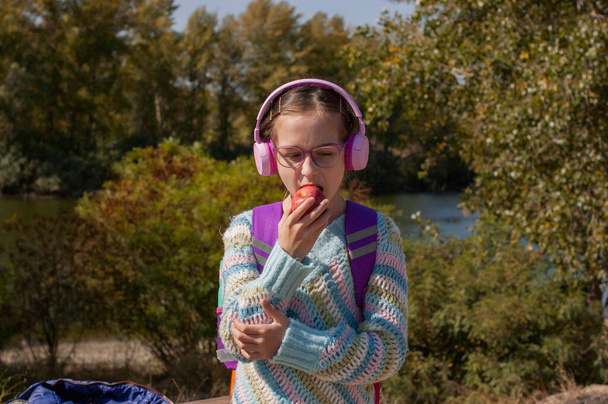 Koulutyttö reppu kuulokkeissa puistossa. Koulutyttö vaaleanpunaisissa kuulokkeissa kuuntelee musiikkia koulun jälkeen. Syksyn koululapsuus. 9-vuotiaan tytön muotokuva - Valokuva, kuva