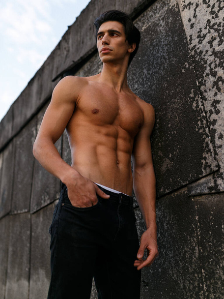 Attractive young man with naked torso near old black wall. Sportive healthy man. Vertical view. - Фото, изображение