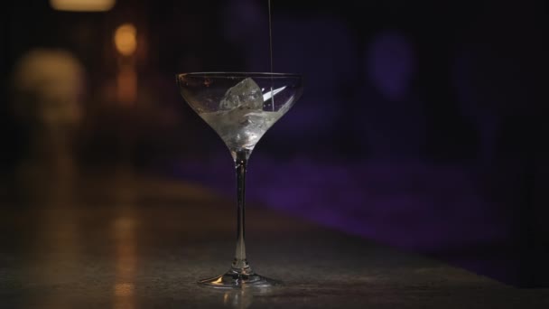 Nahaufnahme des Einfüllens von Alkohol in das Martini-Glas mit Eiswürfel. Archivmaterial. Dickes alkoholisches Getränk wird in transparentes Glas auf dunklem Hintergrund gegossen. - Filmmaterial, Video