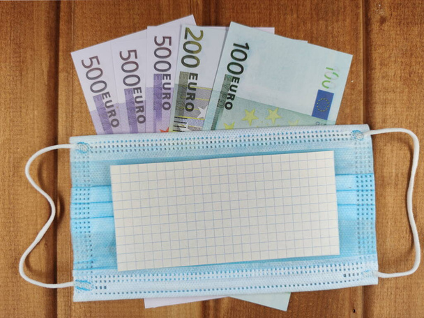 a medical mask are placed on 100, 200 and 500 Euro banknotes and a copy space. background of wooden boards. - Photo, Image