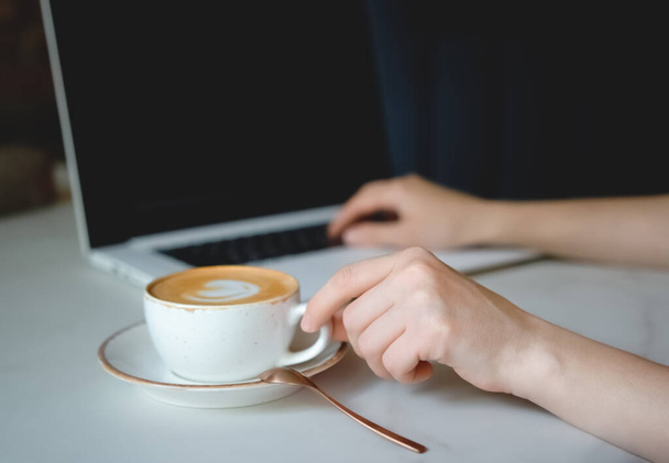 A womans hand picks up a white latte Cup in close-up. Coffee stands on a table on a saucer. Hand print text on a laptop. Cappuccino. Soft focus on the hand. Man using laptop contact. - Fotó, kép