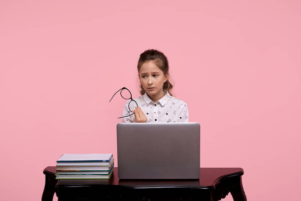 An upset girl sits at a laptop in a white shirt and holds black-framed glasses. - Foto, Imagem